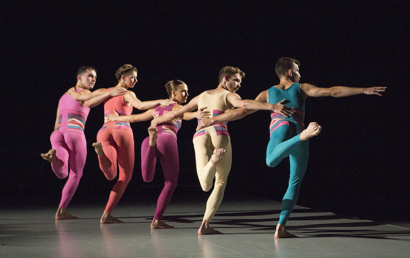 A quintet in sherbet colored unitards link arms with the backs to the audience. They balance on leg and look over their shoulder.
